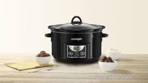 Olla de cocción lenta Opiniones de usuarios, sus ventajas y desventajas y los mejores. en la imagen Una olla de cocción lenta Crockpot negra colocada sobre una mesa de madera clara, acompañada de tres pequeños cuencos blancos con comida y un paño amarillo con utensilios. El fondo es minimalista con tonos beige. En la parte inferior derecha de la imagen aparece la marca "SoyInsider.com".